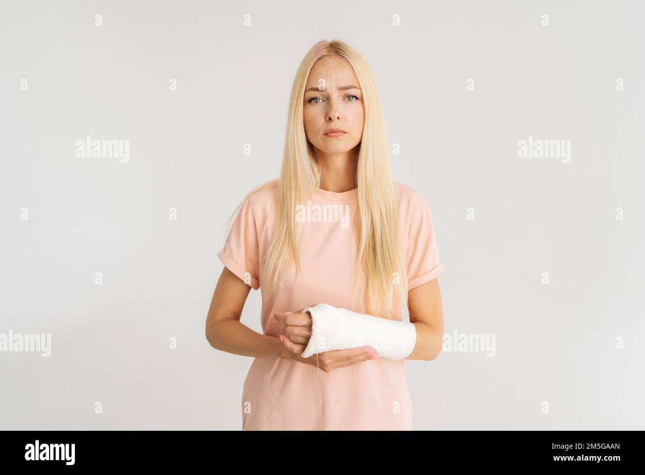 Studio portrait de malheureuse blessée jeune femme blonde avec bras cassé enveloppé dans le bandage de plâtre regardant la caméra, debout sur blanc isolé Banque D'Images