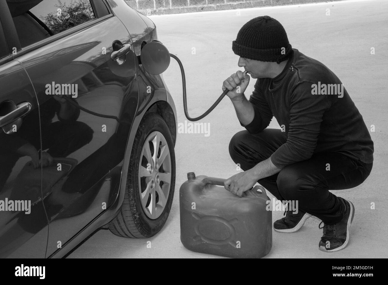 Image d'un voleur vêtu de noir qui aspire l'essence d'un réservoir de voiture avec un tuyau en caoutchouc. Référence à l'augmentation du vol de carburant en raison d'un coût excessif. Banque D'Images