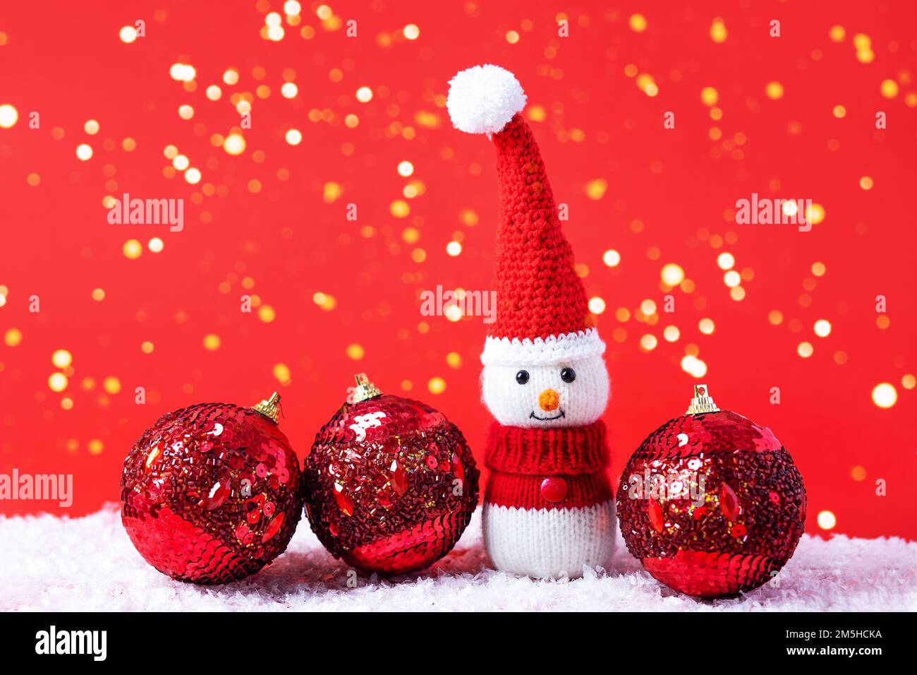 Bonhomme de neige tricoté dans un chapeau de Noël rouge et un chandail avec trois boules rouges sur un fond rouge avec des lumières de Noël dorées. Joyeux Noël et nouvel an Banque D'Images