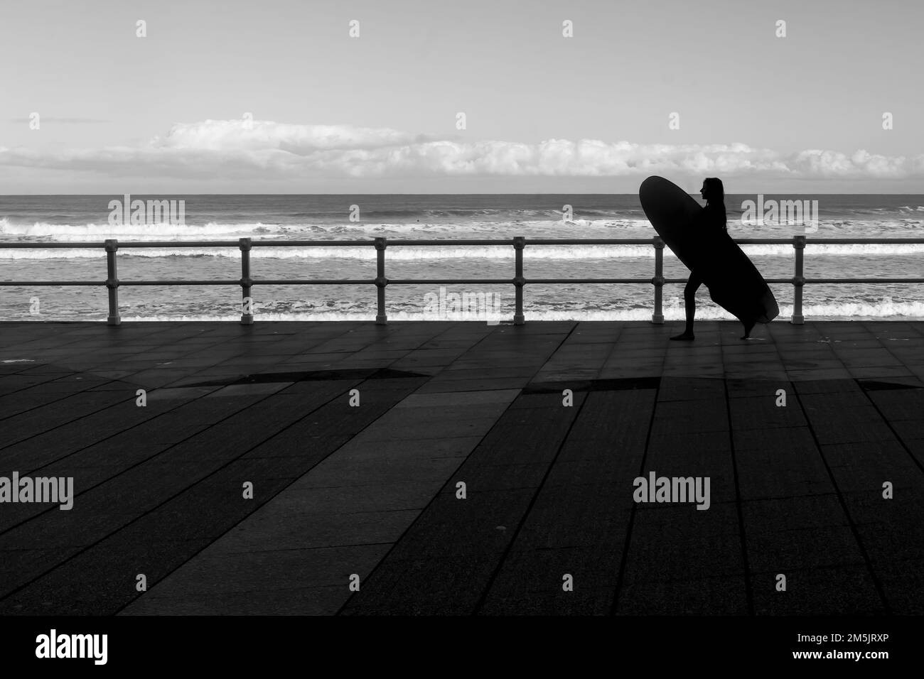 Vue sur la silhouette d'une femme tenant une planche de surf et marchant le long de la promenade de Gijon Asturias, Espagne. Banque D'Images