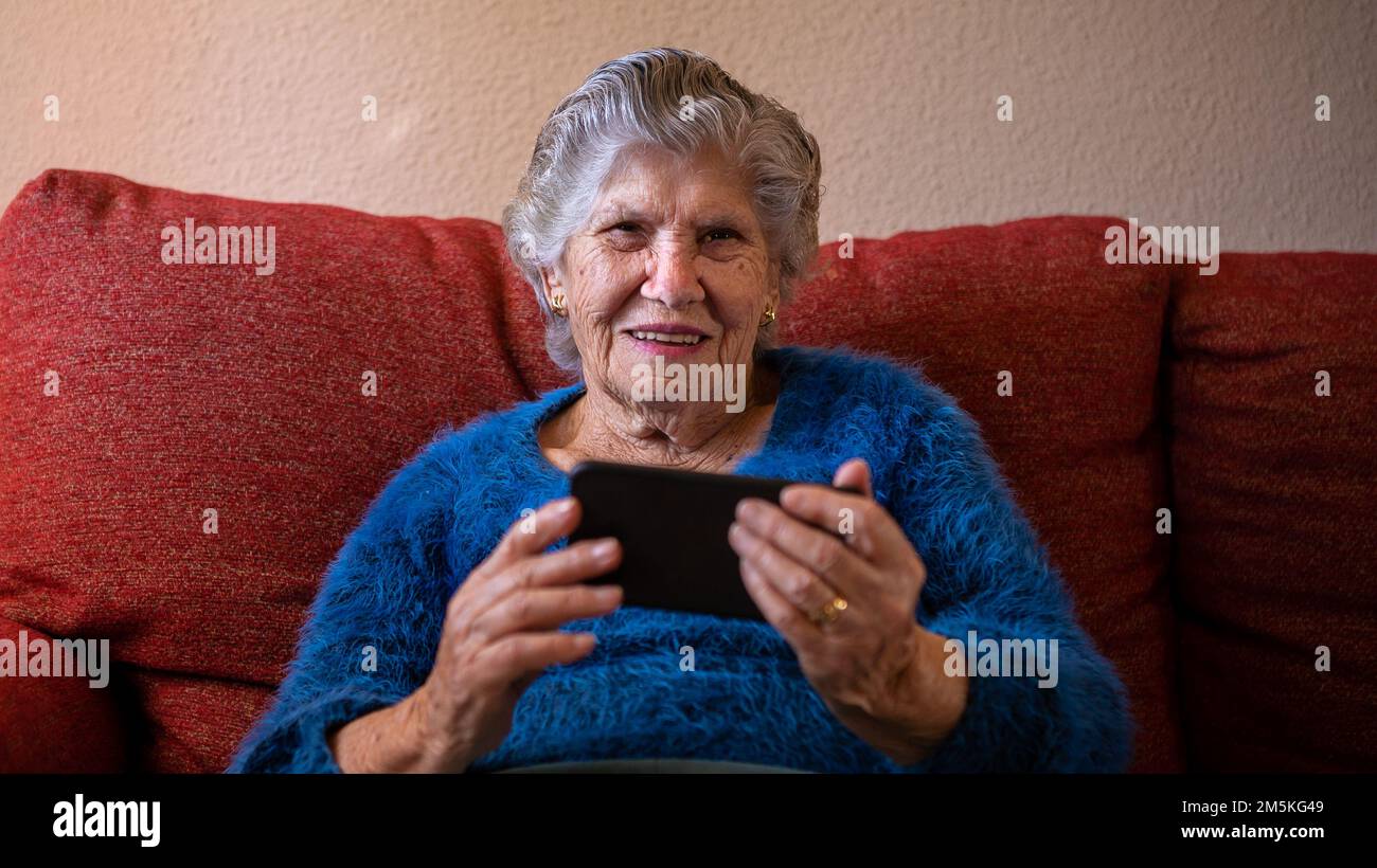 Femme âgée regardant l'appareil photo tout en utilisant un téléphone mobile assis sur un canapé maison. Une grand-mère souriante tient un smartphone. Ancien grand-mère exploitant un Banque D'Images