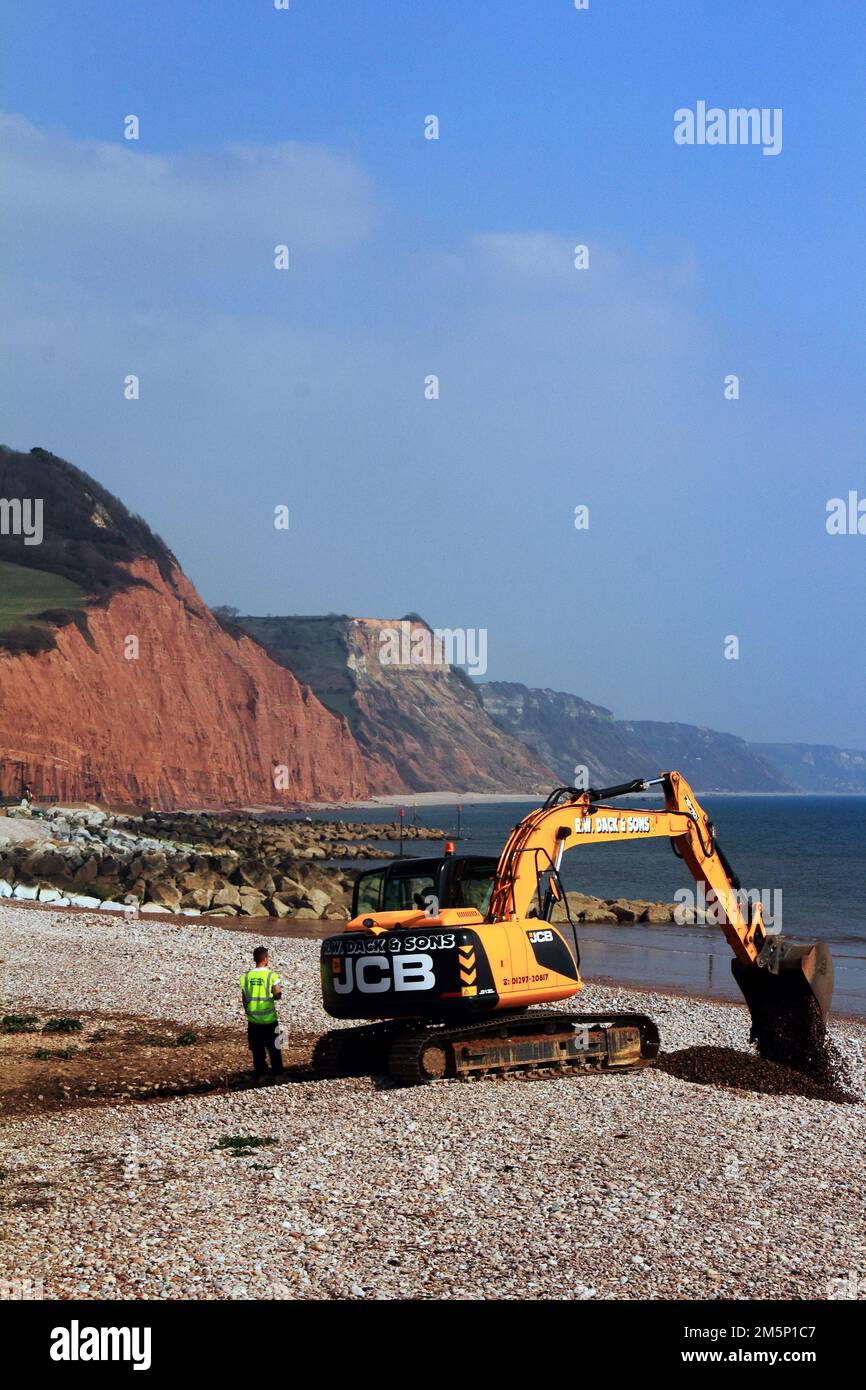 Un creuseur et un opérateur JCB sur la plage de galets à l'extrémité est de la plage de Sidmouth avec la falaise de Salcombe Hill en grès rouge et la côte jurassique Banque D'Images