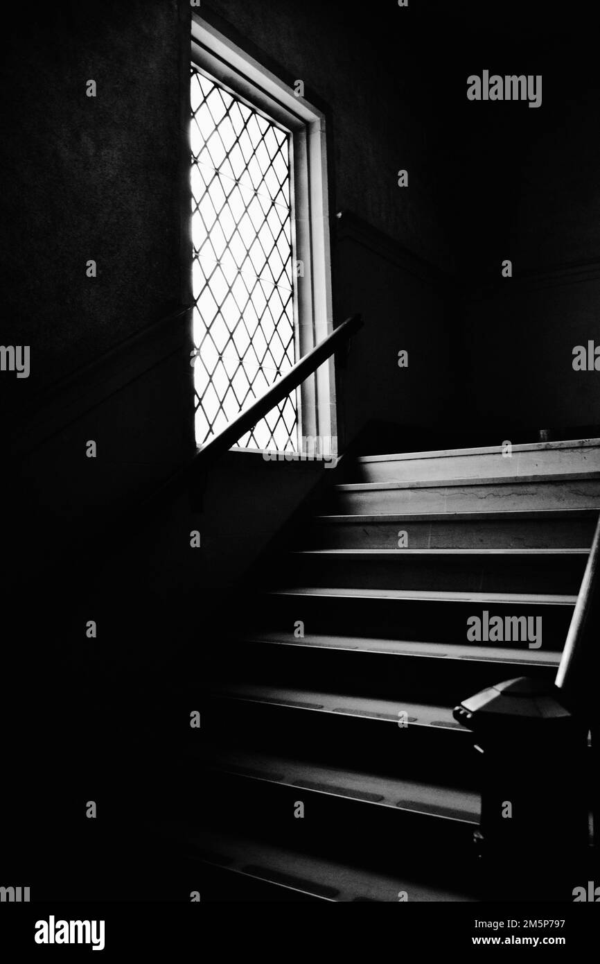 Une échelle de gris verticale des escaliers et une fenêtre avec des rampes dans un bâtiment sombre Banque D'Images