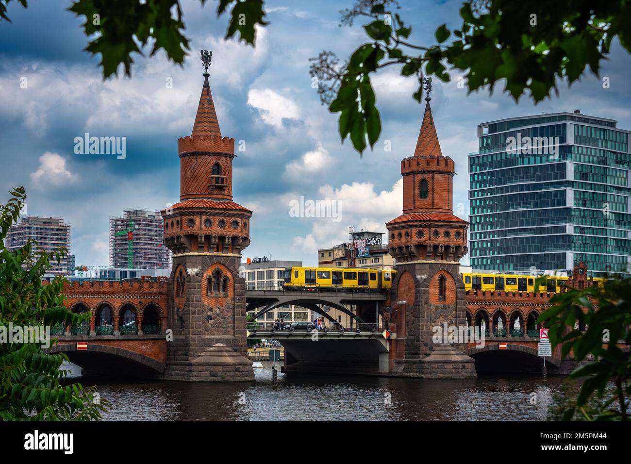 Berlin, Allemagne - 25 juin 2022 : symbole de Berlin : le pont Oberbaum, entouré de branches d'arbres tandis qu'un tramway jaune vif traverse Banque D'Images