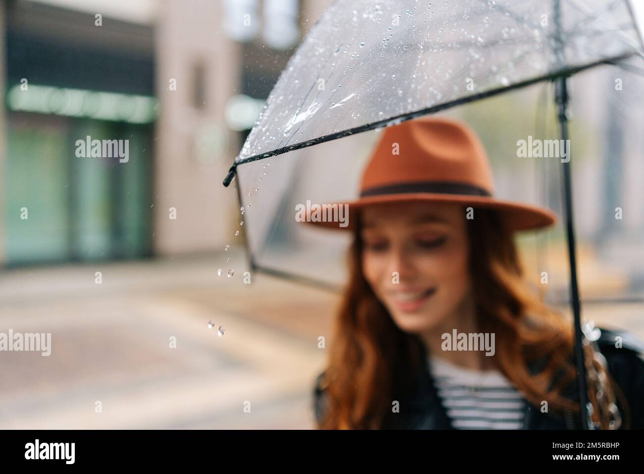 Objectif sélectif de charmante jeune femme portant un chapeau élégant debout avec un parapluie transparent sur la rue de la ville profitant du temps pluvieux dehors. Banque D'Images