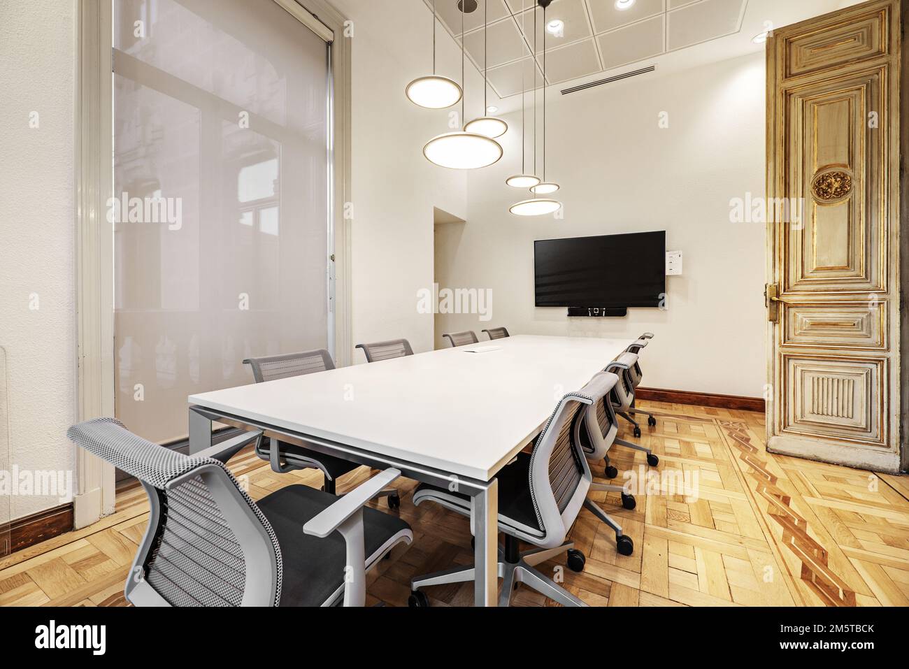 Salle de réunion d'un imposant bâtiment d'époque avec une longue table en bois blanc avec chaises pivotantes grises, parquet et lampes design Banque D'Images