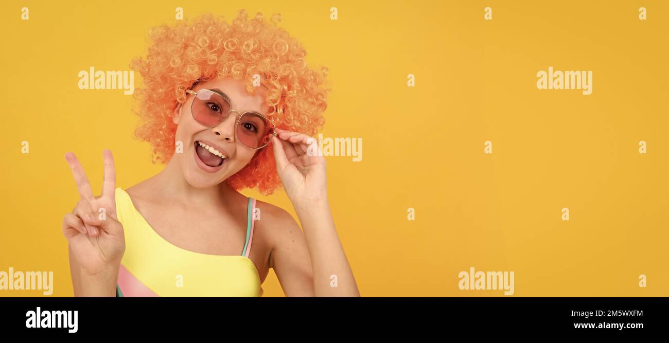 joyeux enfant dans des lunettes de soleil et un maillot de bain portant des cheveux de perruque orange montrant le geste de paix, la paix. Adolescent drôle sur la fête, affiche en-tête de bannière Banque D'Images