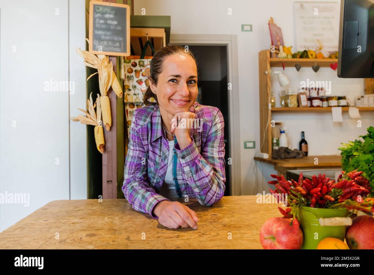 Belle femme greengrocer travaillant dans la boutique penchée sur le comptoir et entourée de fruits et légumes biologiques - Portrait de la jeune femme entrepreneur Banque D'Images