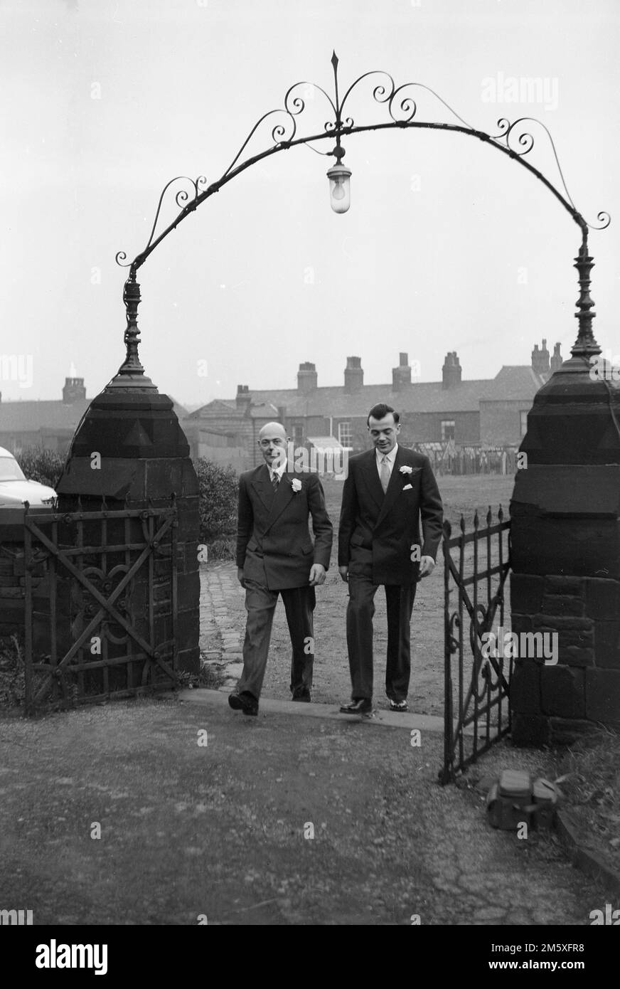 Années 1950 historique, un marié et meilleur homme marchant sous une arche décorative en métal à l'entrée extérieure du terrain de l'église, Angleterre, Royaume-Uni. Banque D'Images