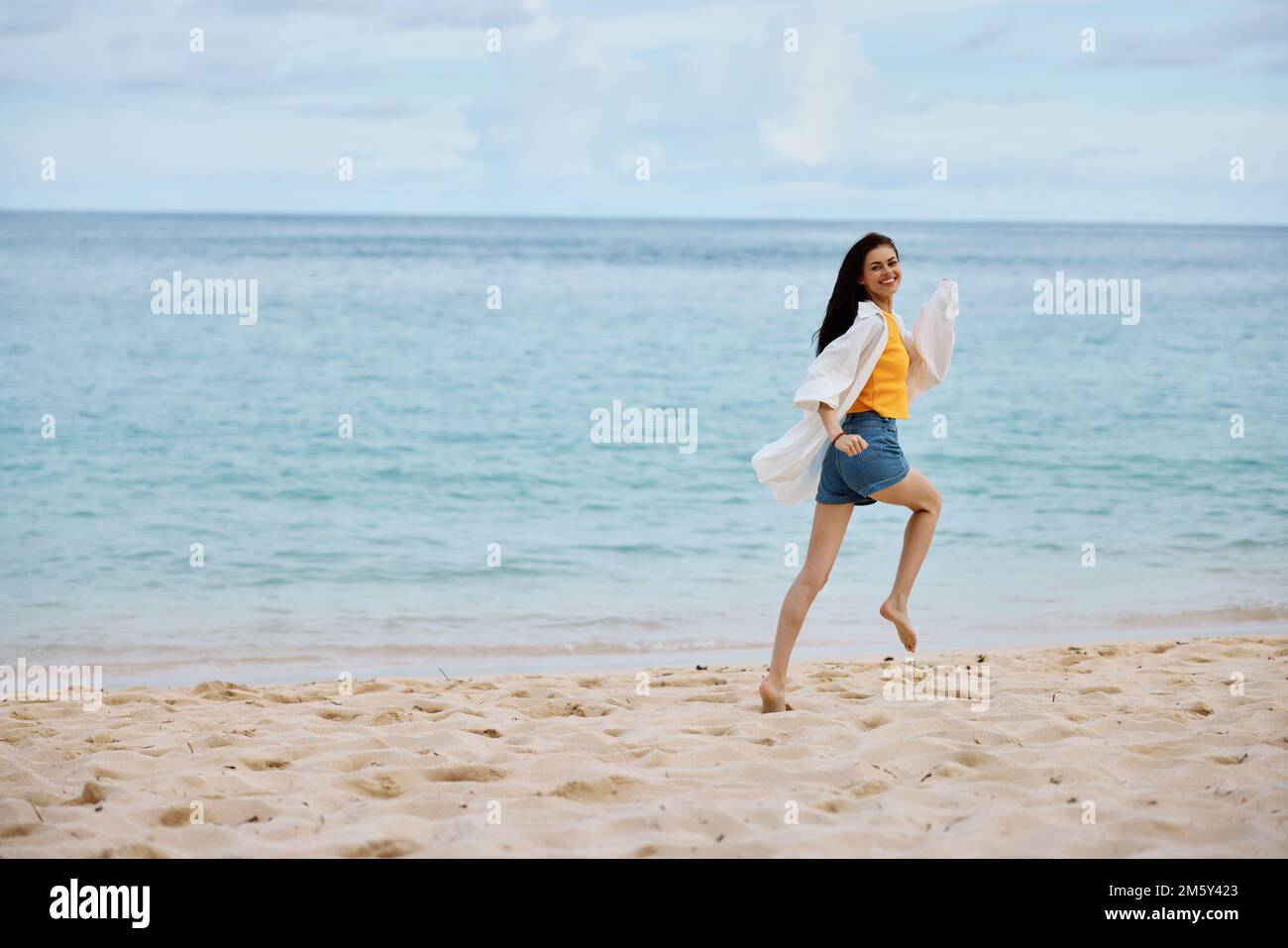 Femme de sport court le long de la plage en vêtements d'été sur le sable dans un t-shirt jaune et short en denim blanc shirt vol cheveux vue sur l'océan, plage Banque D'Images