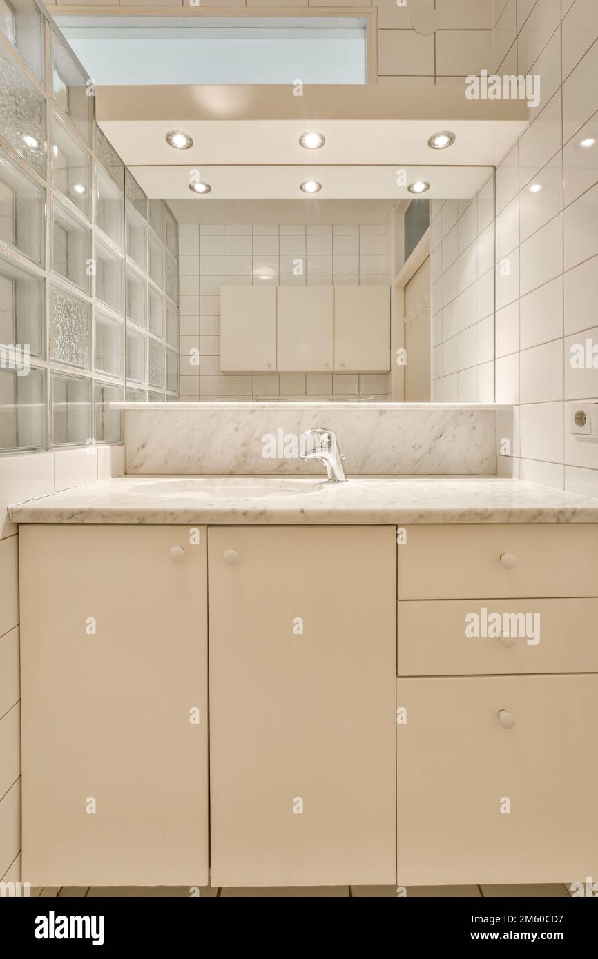 une salle de bains avec des armoires blanches et des plans de travail en marbre sur le meuble-lavabo en face du lavabo est éclairée par des lumières encastrées Banque D'Images