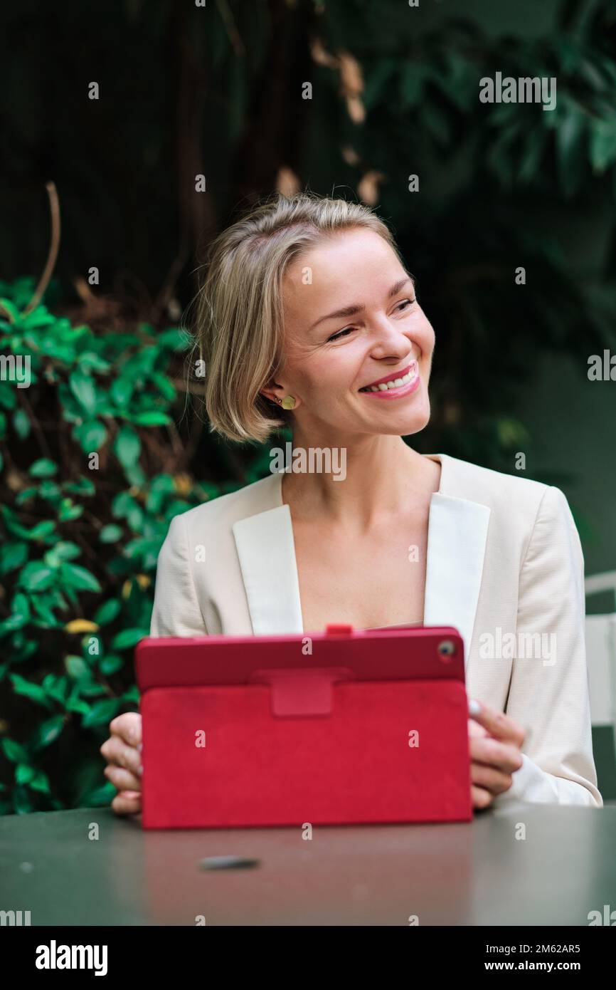 Femme regardant loin et souriant tout en utilisant une tablette numérique assis sur une table à l'extérieur. Banque D'Images