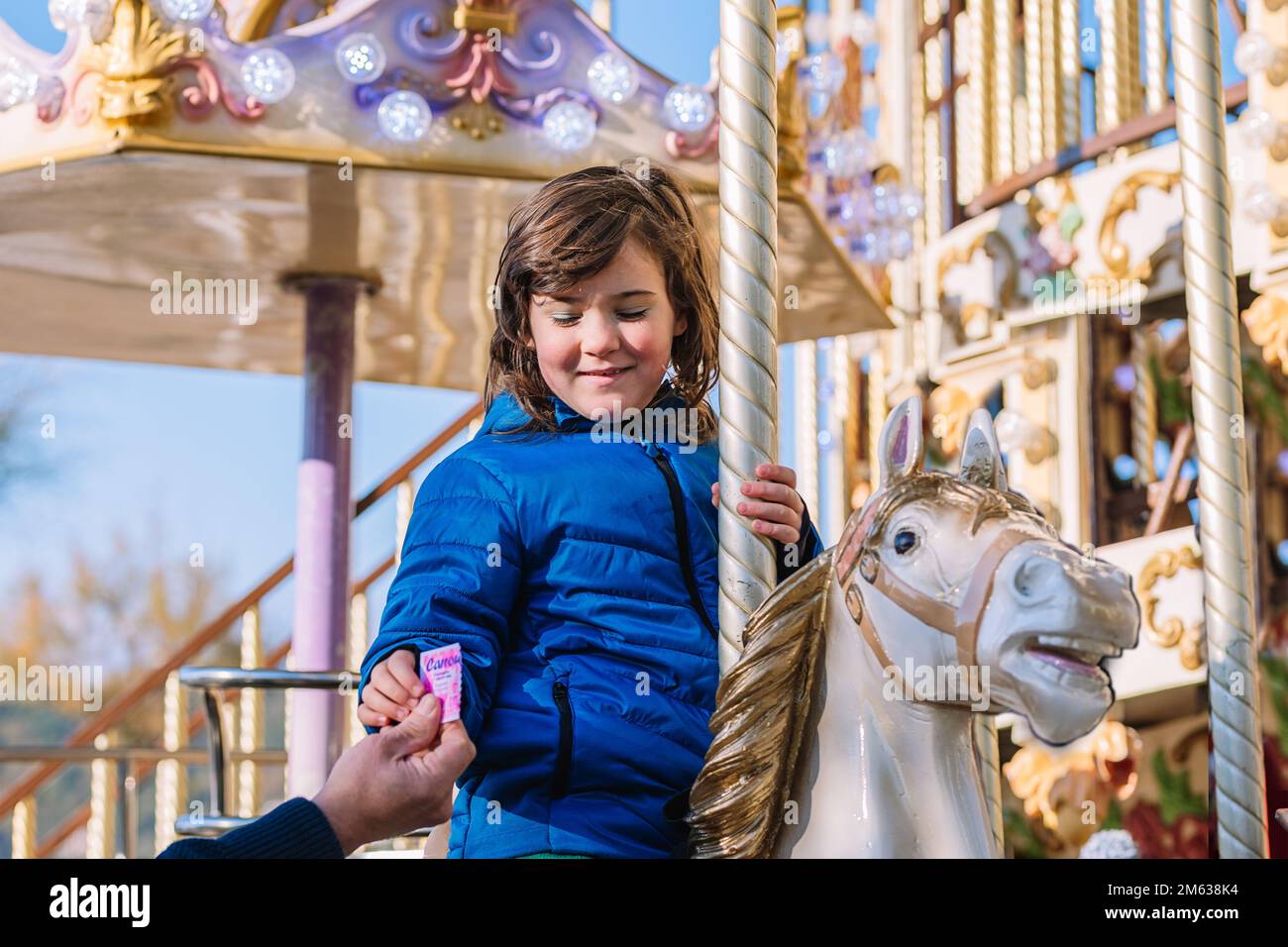 Petit angle de fille gaie dans un manteau bleu souriant et donnant le billet à l'ouvrier tout en étant assis sur le joyeux aller cheval rond dans la journée à Donostia San Sebastian Banque D'Images