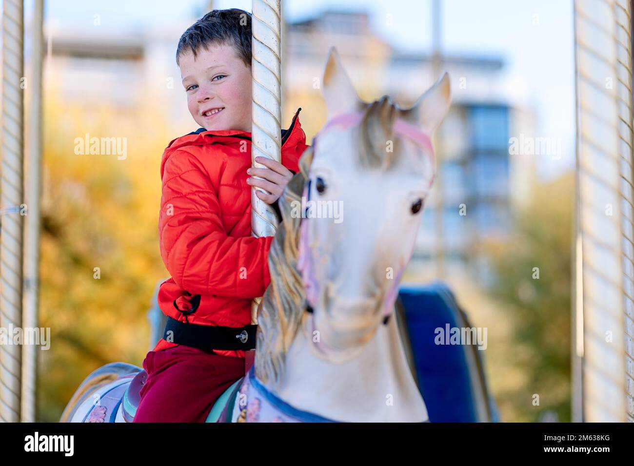 Joyeux garçon en vêtements d'extérieur souriant et assis regardant l'appareil photo à cheval tout en faisant du carrousel le jour ensoleillé sur le champ de foire de Donostia San Sebastian, SPAI Banque D'Images