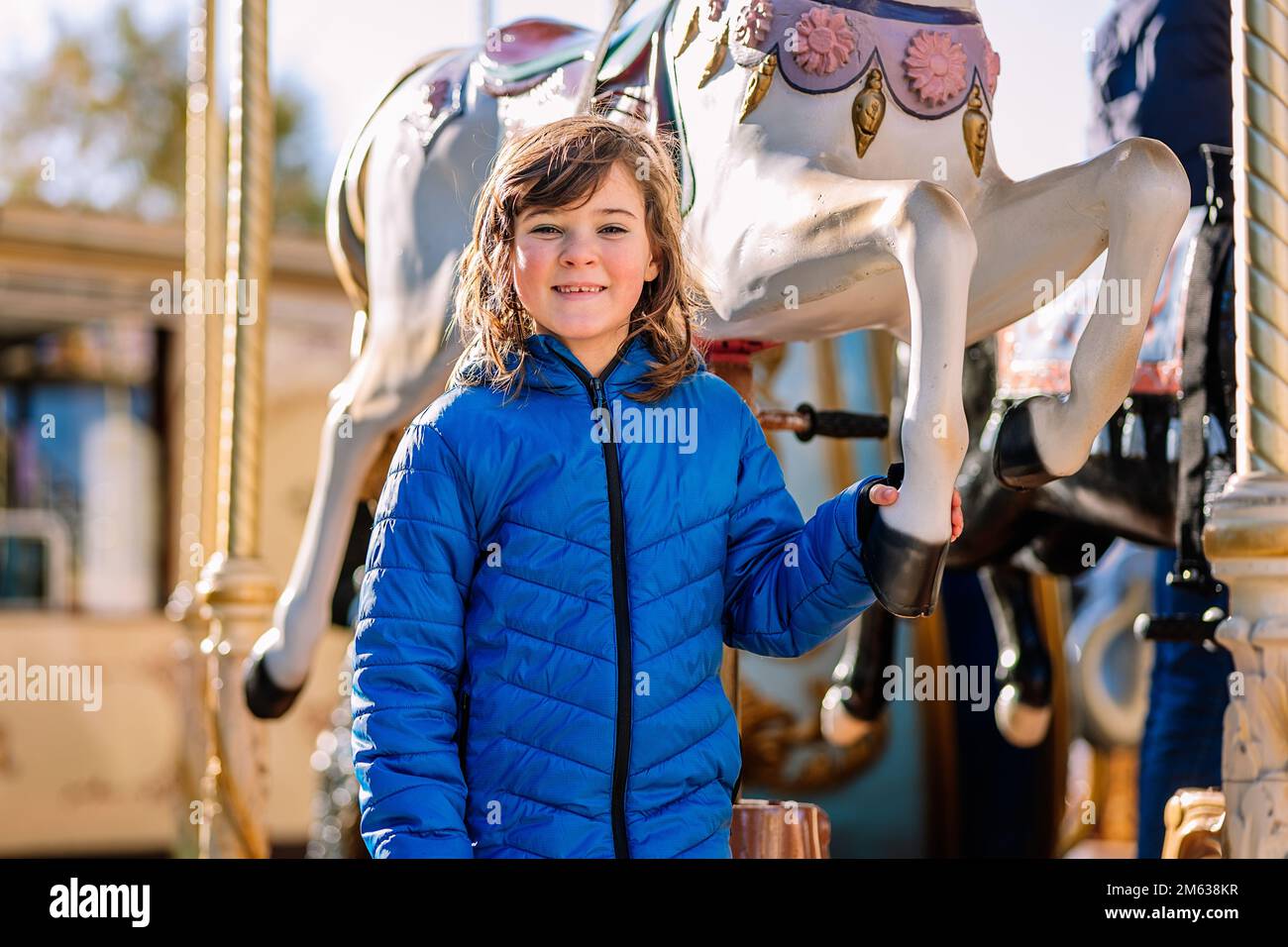 Bonne fille en manteau bleu touchant la jambe du cheval de carrousel et regardant l'appareil photo avec le sourire tout en passant une journée de week-end sur le terrain d'exposition de Donostia San Sebas Banque D'Images