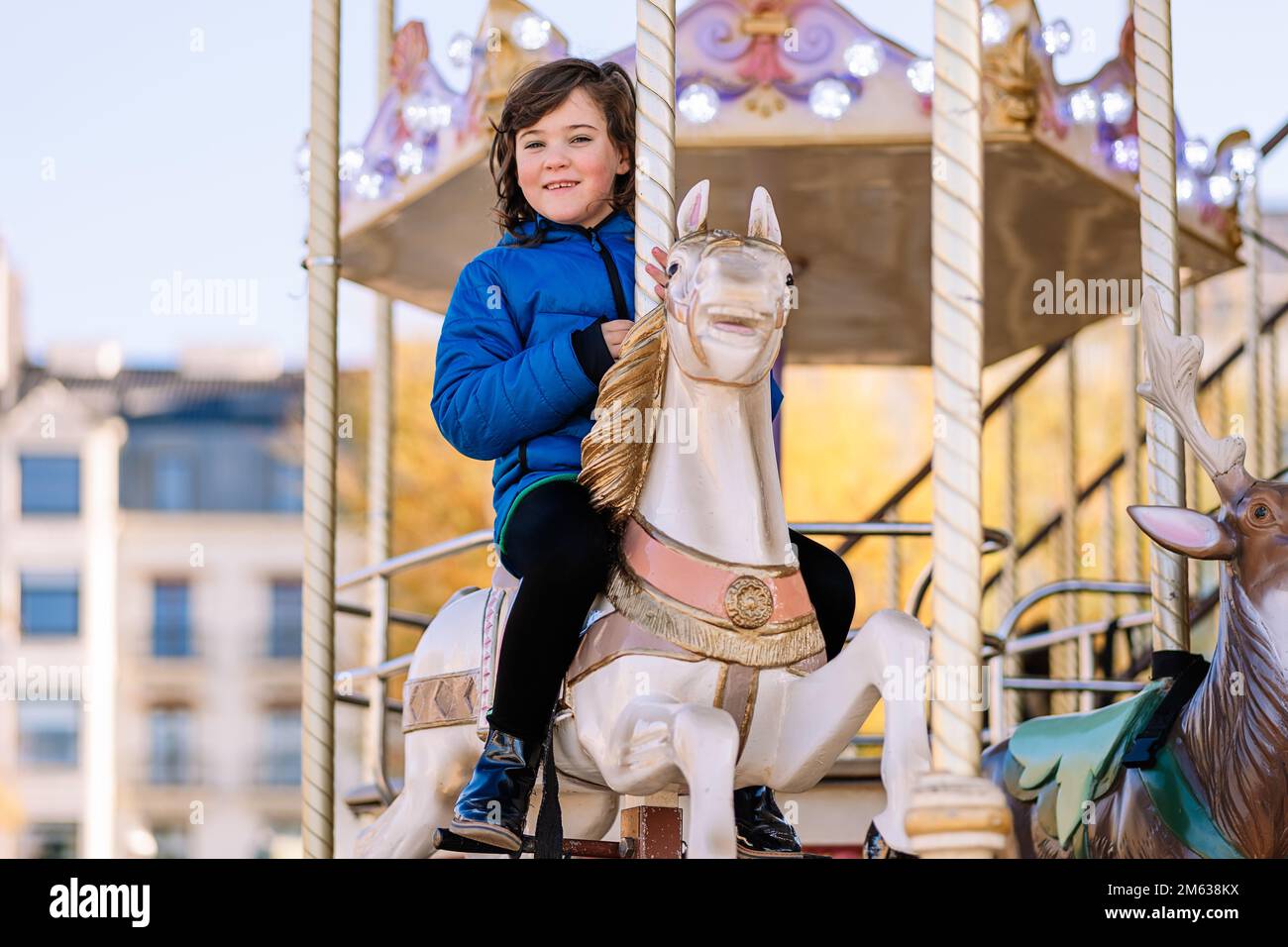 Bonne fille en vêtements d'extérieur souriant et assis regardant l'appareil photo à cheval tout en faisant du carrousel à cheval le jour ensoleillé sur le parc d'expositions de Donostia San Sebastian, Spa Banque D'Images