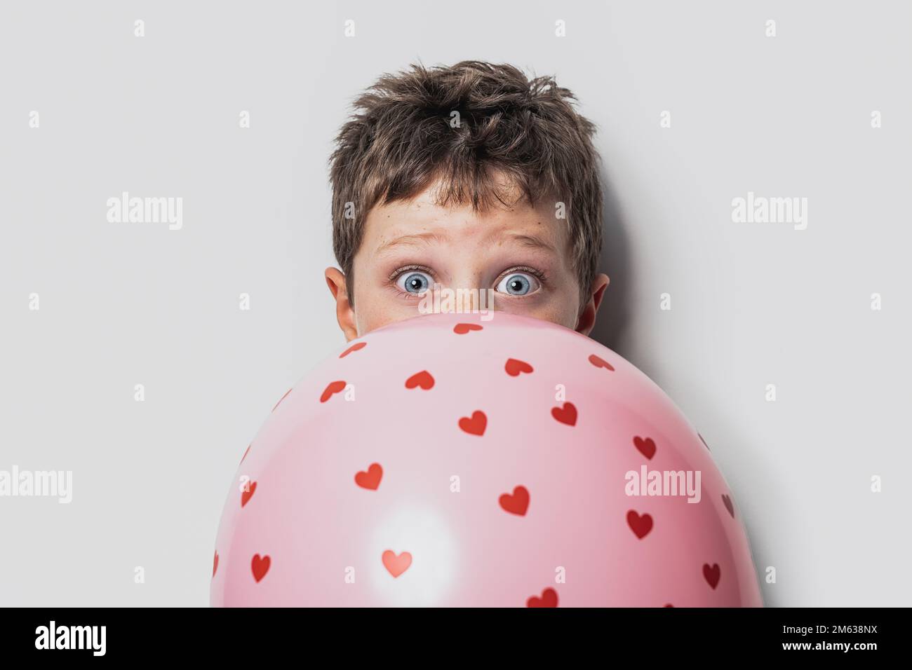 Un joli garçon aux yeux bleus cache le visage derrière un ballon rose avec  des coeurs et regarde la caméra contre le mur gris le jour de la  Saint-Valentin Photo Stock -