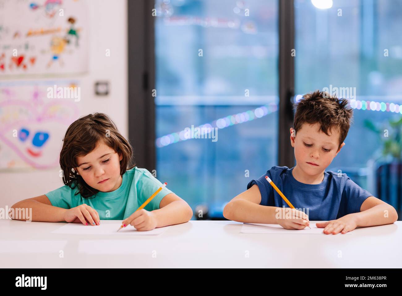 Frère et sœur concentrés en t-shirts bleus assis et table et esquissant le soir à la maison Banque D'Images Frère et sœur concentrés en t-shirts bleus assis et table et esquissant le soir à la maison Banque D'Images