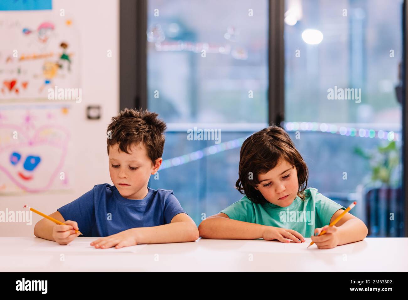 Frère et sœur concentrés en t-shirts bleus assis et table et esquissant le soir à la maison Banque D'Images Frère et sœur concentrés en t-shirts bleus assis et table et esquissant le soir à la maison Banque D'Images