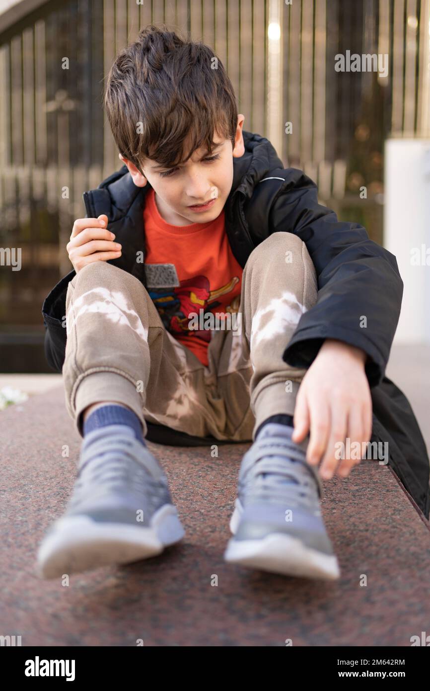 Inquiet jeune garçon douleur visage mains joint tenir la jambe douleur en raison de la cheville cassée dans la rue extérieure Banque D'Images