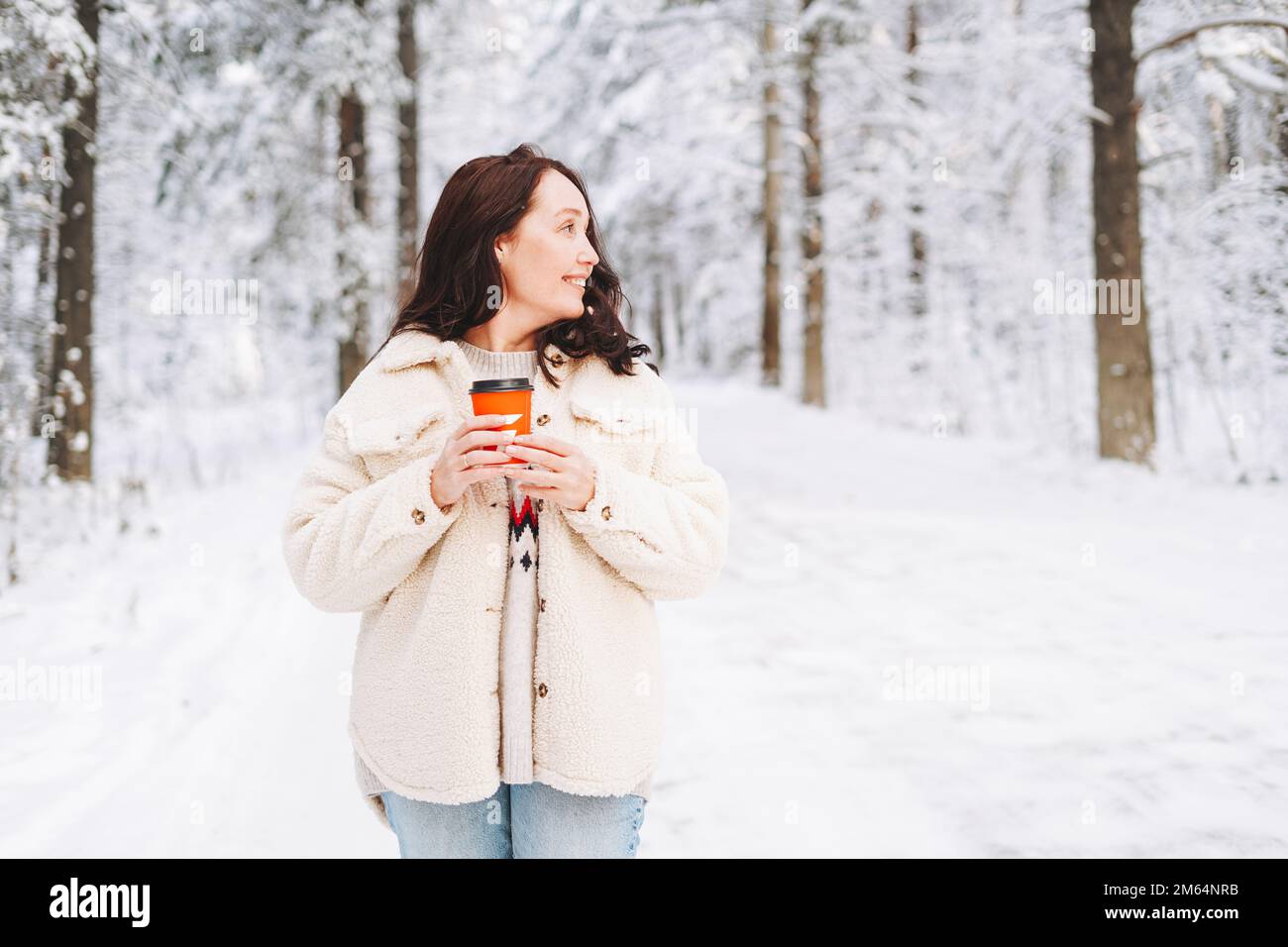 Femme souriante aux cheveux foncés dans des vêtements d'hiver avec une tasse de papier de café entre les mains sur fond de forêt enneigée d'hiver Banque D'Images