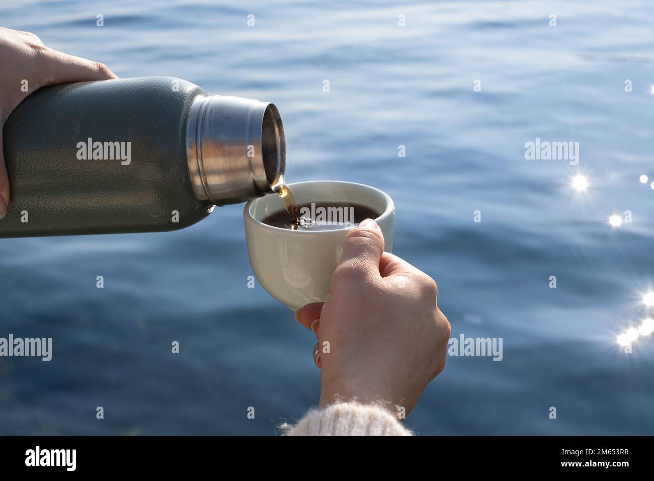 la main de femme verse la boisson chaude des thermos dans le verre. Tout en versant une boisson d'un thermos dans un verre, la mer est en arrière-plan. Banque D'Images