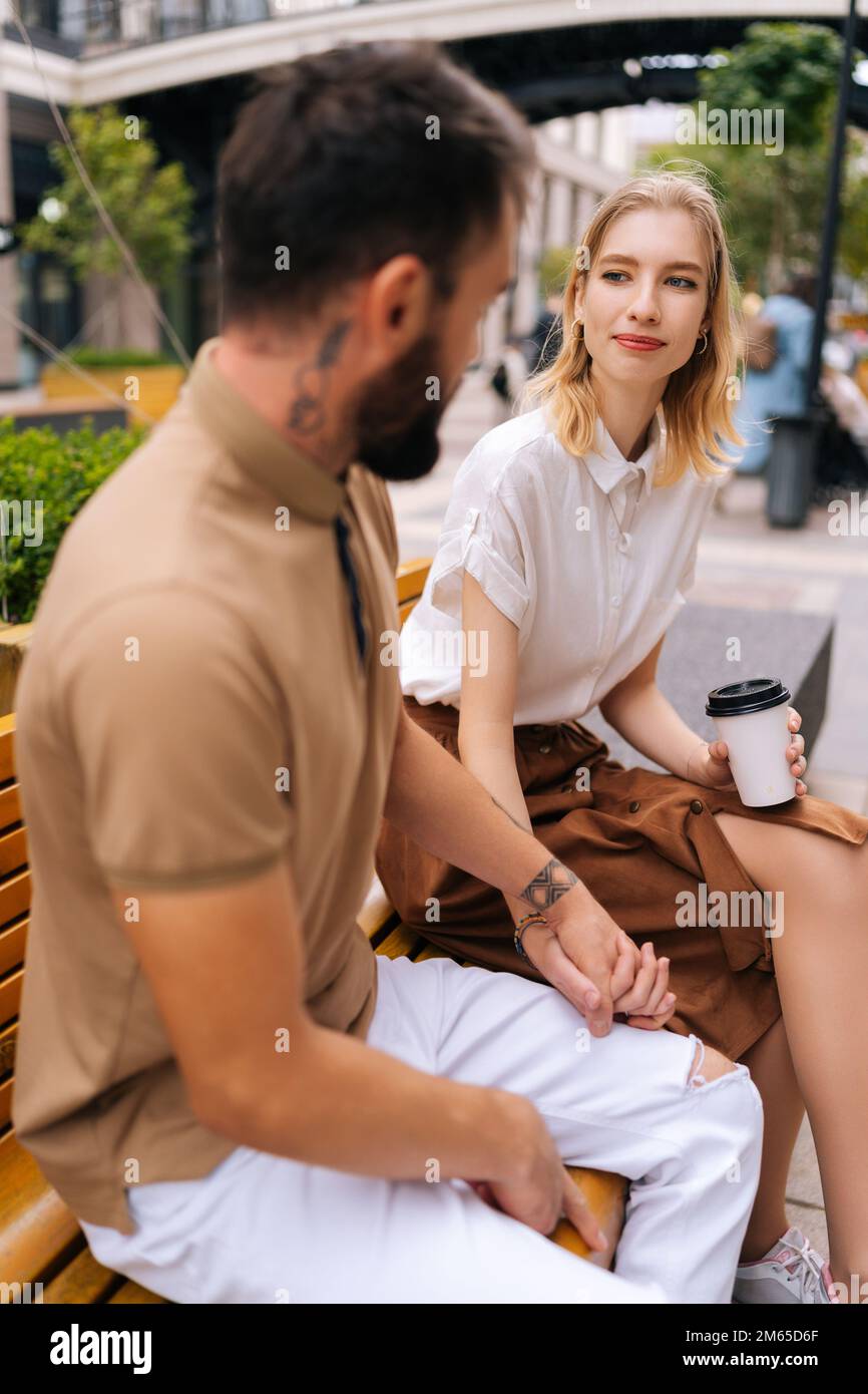 Vue verticale sur le côté d'une jeune femme élégante aux cheveux blonds parlant avec un homme tatoué barbu assis sur un banc avec un café à emporter dans le parc. Banque D'Images