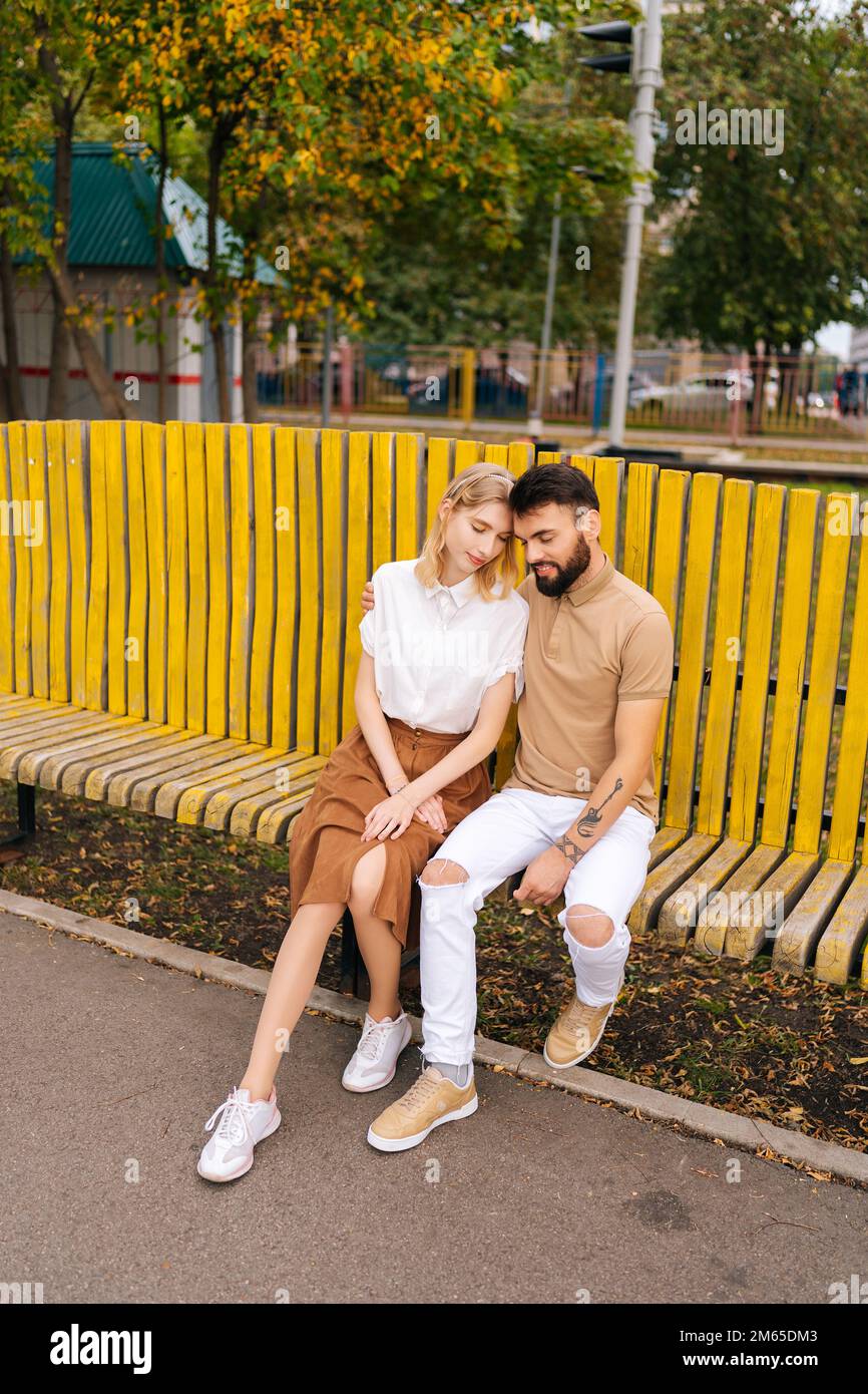 Portrait vertical de jeune couple aimant se reposant dans le parc de la ville embrassant avec les yeux fermés sur le banc en été ensoleillé jour. Banque D'Images