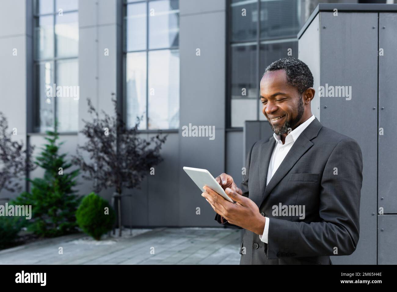 Homme d'affaires afro-américain prospère souriant et utilisant un Tablet PC, homme d'affaires à l'extérieur d'un immeuble de bureau moderne marchant. Banque D'Images