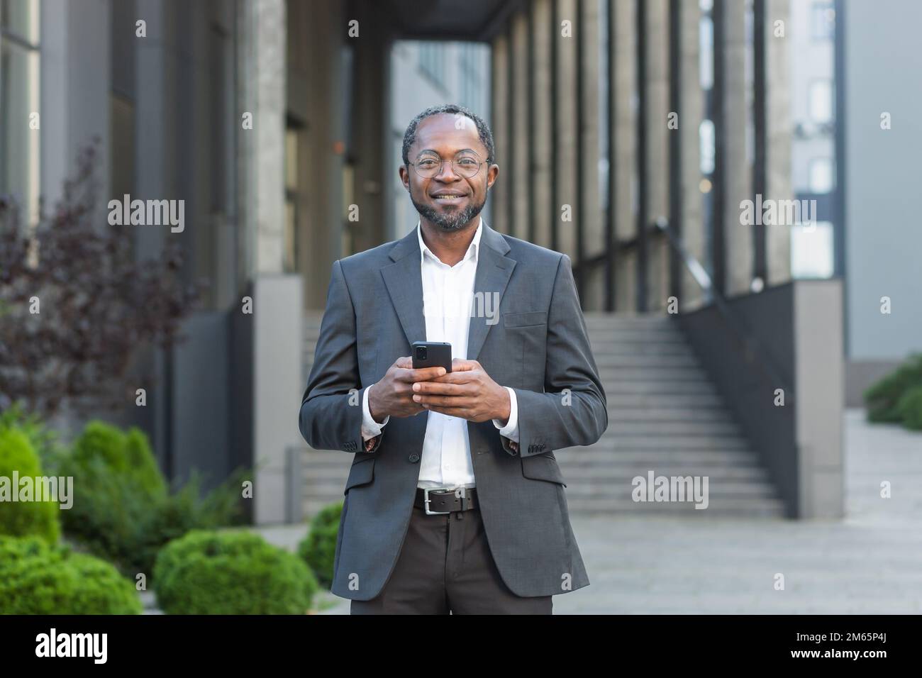 Portrait d'homme d'affaires afro-américain mature, homme d'affaires senior à l'extérieur du bâtiment de bureau tenant le téléphone entre les mains souriant et regardant l'appareil photo. Banque D'Images