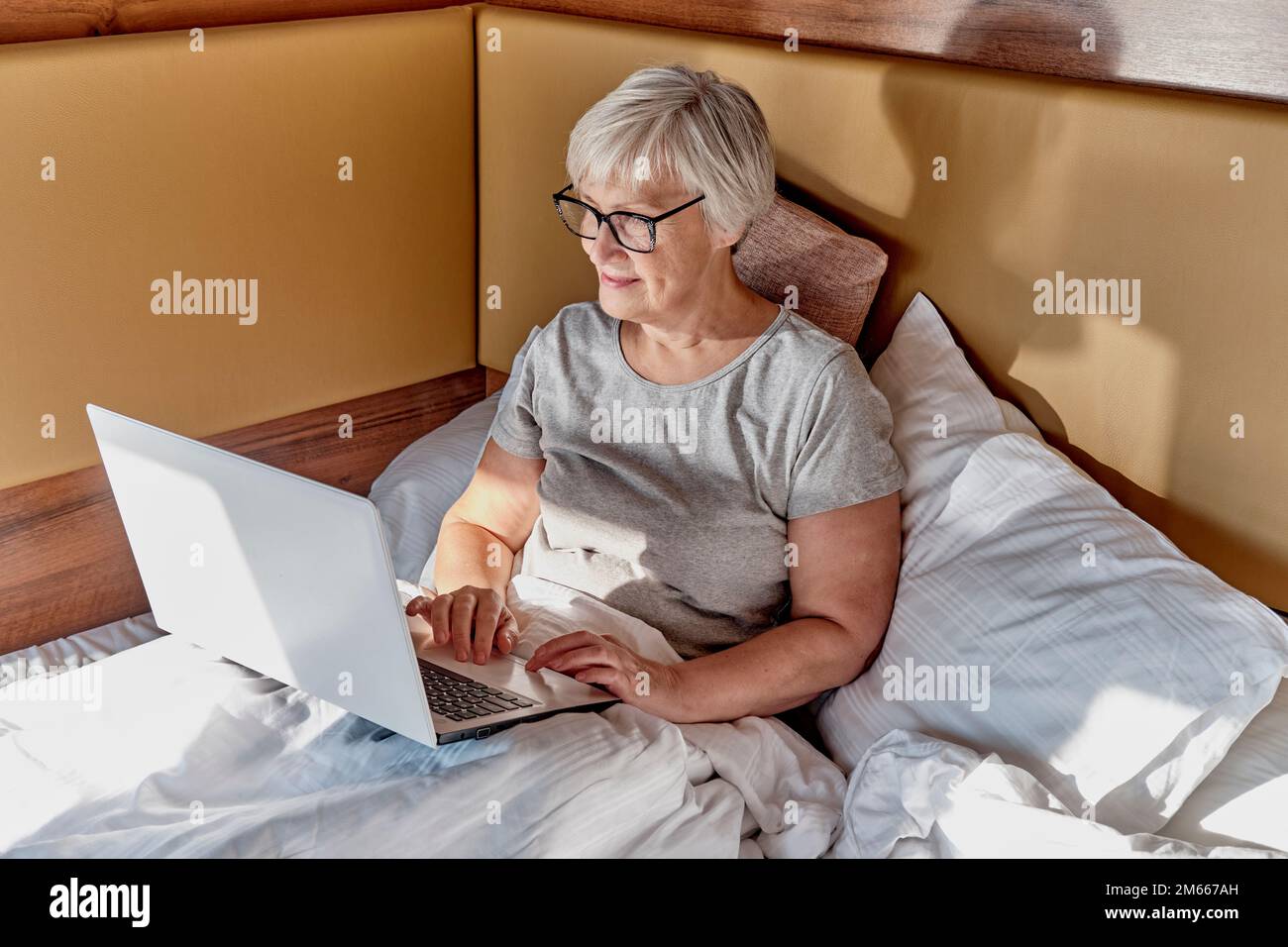 Femme aînée souriante aux cheveux gris courts, utilisant un ordinateur portable dans le lit. À la maison, à l'hôtel, en camping. Travailler en ligne, utiliser des services en ligne, en acheter Banque D'Images