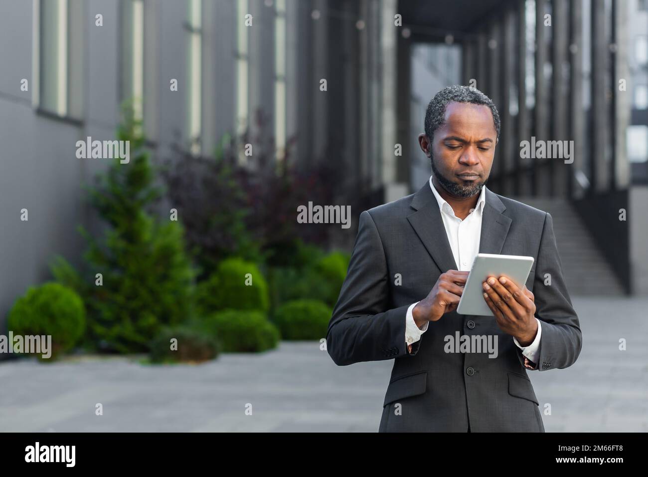Un homme d'affaires afro-américain sérieux qui utilise une tablette, un homme d'affaires suit des informations mûres en ligne debout à l'extérieur de l'immeuble de bureaux. Banque D'Images