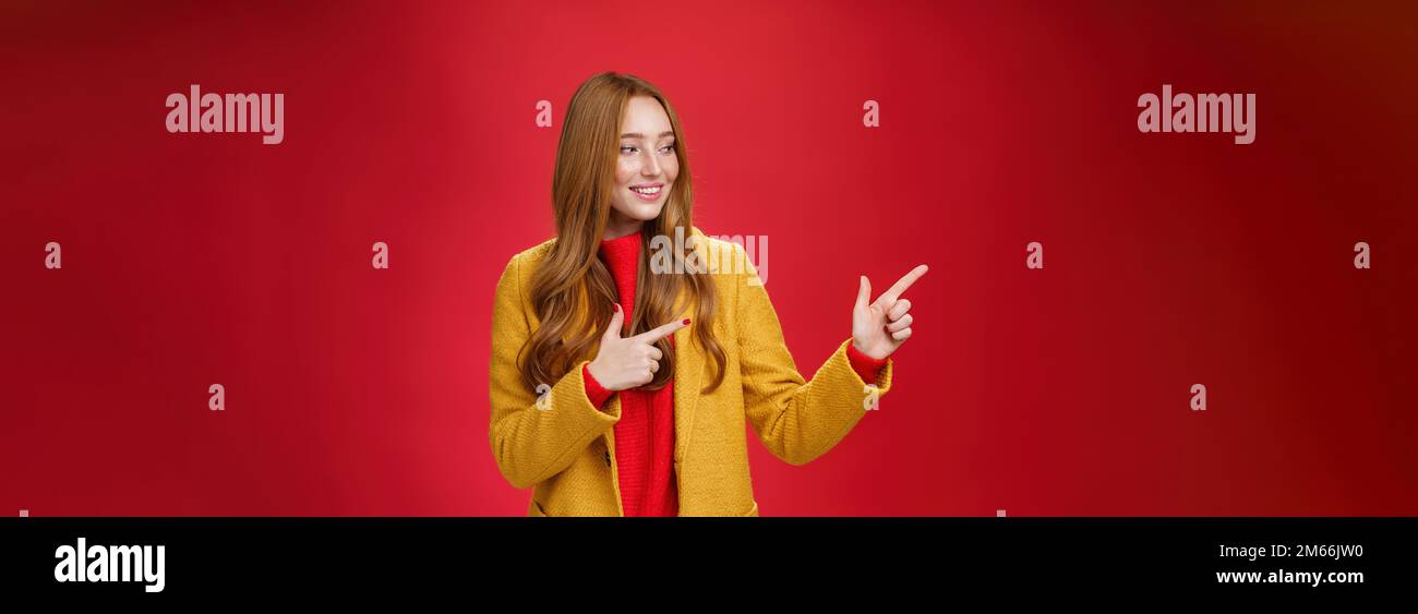 Portrait de curieuse et heureuse heureuse heureuse belle femme à tête rouge dans le manteau jaune pointant et regardant à gauche avec heureux intrigué sourire observer Banque D'Images