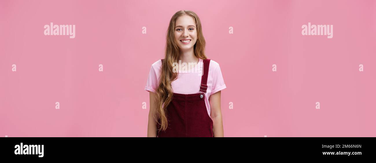 Portrait de charmante petite fille mince avec de longs cheveux justes dans des combinaisons en velours côtelé souriant debout joyeusement debout avec sourire positif de sourire posé contre le rose Banque D'Images