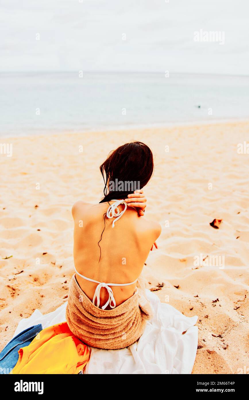 Une femme est assise avec elle dos à la caméra après avoir nagé dans l'océan dans un maillot de bain sur une serviette sur le sable et regarde la mer et les nuages, l'été Banque D'Images