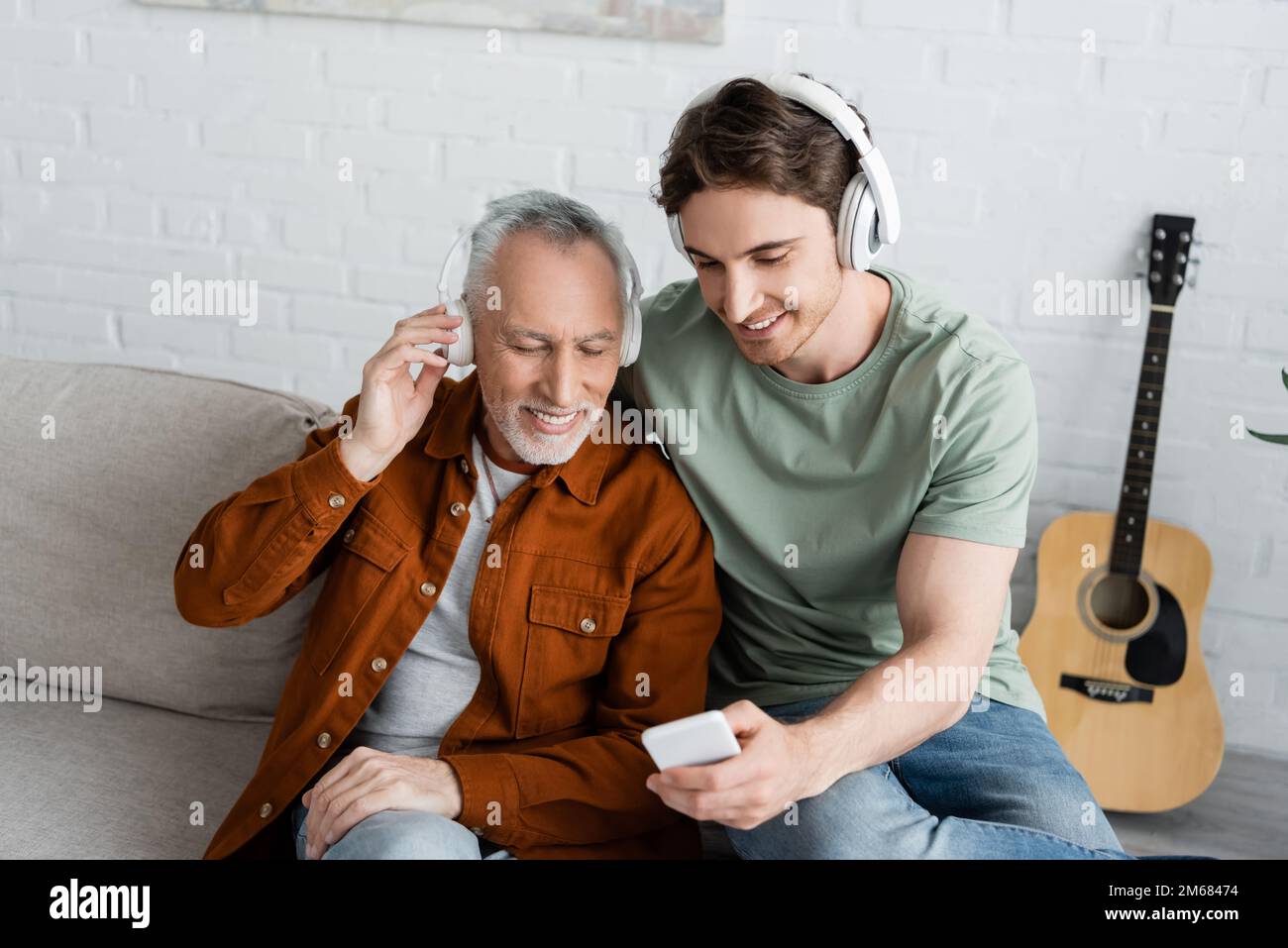 homme souriant montrant un smartphone à un homme aux cheveux gris heureux dans un casque sans fil tout en écoutant de la musique à la maison, image de stock Banque D'Images