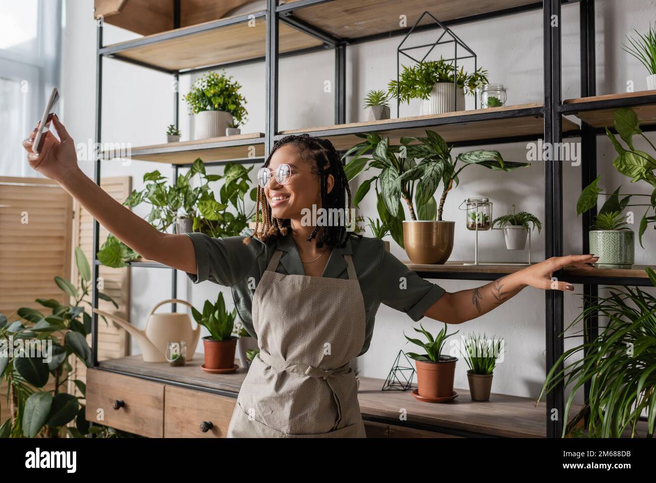 fleuriste afro-américain souriant avec téléphone mobile debout près du rack avec des plantes en pot pendant la commande en ligne, image de stock Banque D'Images