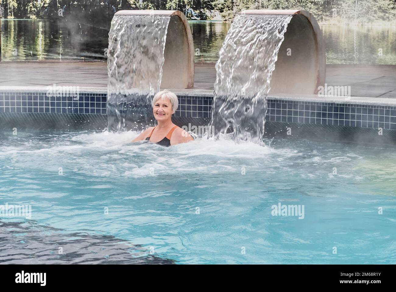 Femme aînée souriante heureuse avec cheveux gris courts regardant la caméra dans la piscine thermale extérieure avec hydromassage.Active Seniors concept. Banque D'Images