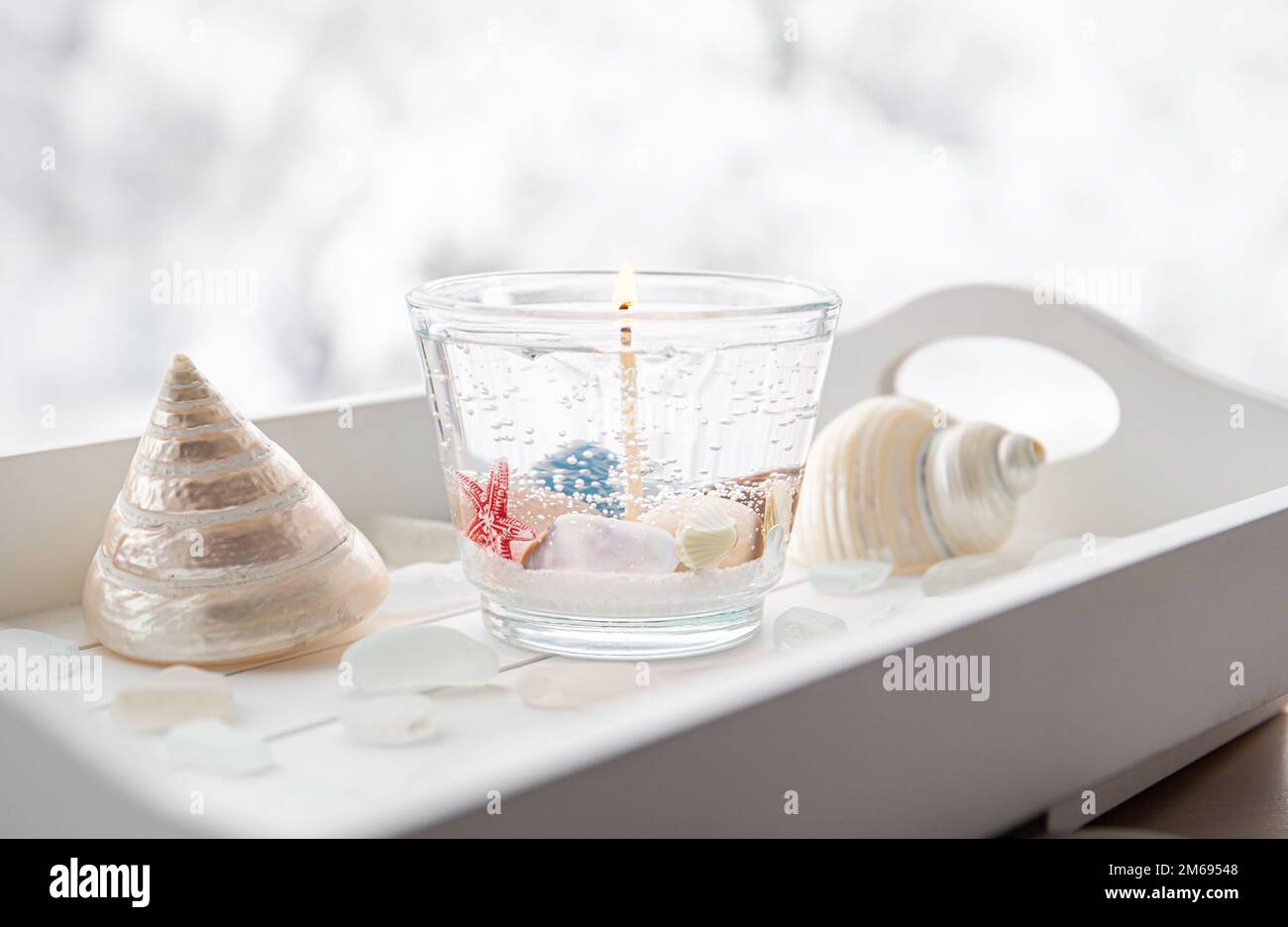 Bougie de cire de gel transparente sous-marine à la main à l'intérieur d'une tasse transparente. Belle bougie sur plateau en bois blanc avec verre de mer et coquillages sur la fenêtre. Banque D'Images
