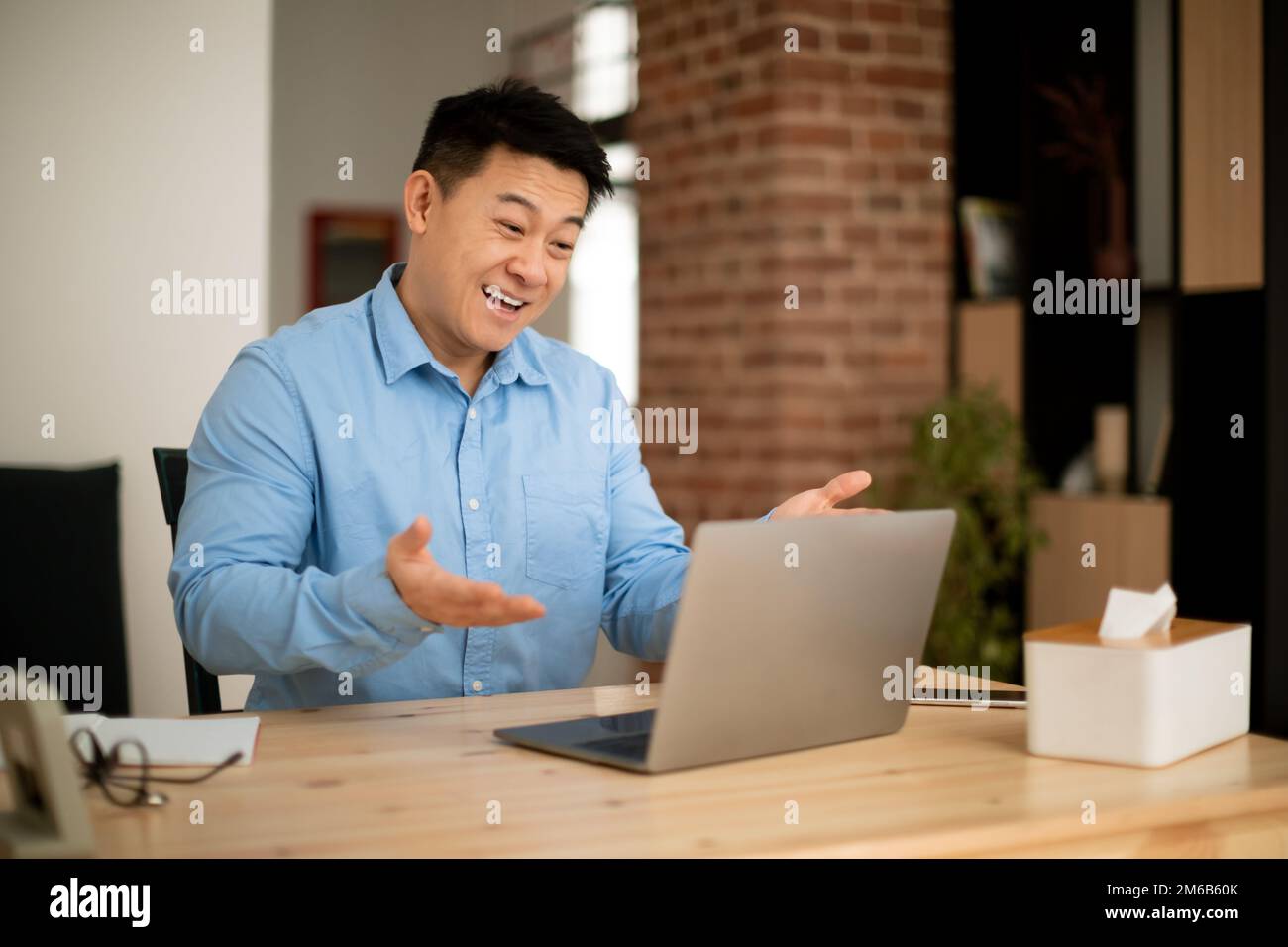Homme asiatique mature et enthousiaste utilisant un ordinateur portable moderne, des appels vidéo et des gestations à la webcam, assis sur le lieu de travail à la maison Banque D'Images