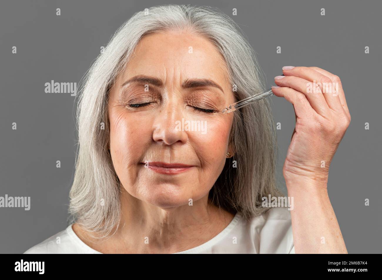 Sérieuse assez vieille femme caucasienne avec les yeux fermés applique l'huile ou le sérum au visage, isolé sur fond gris Banque D'Images