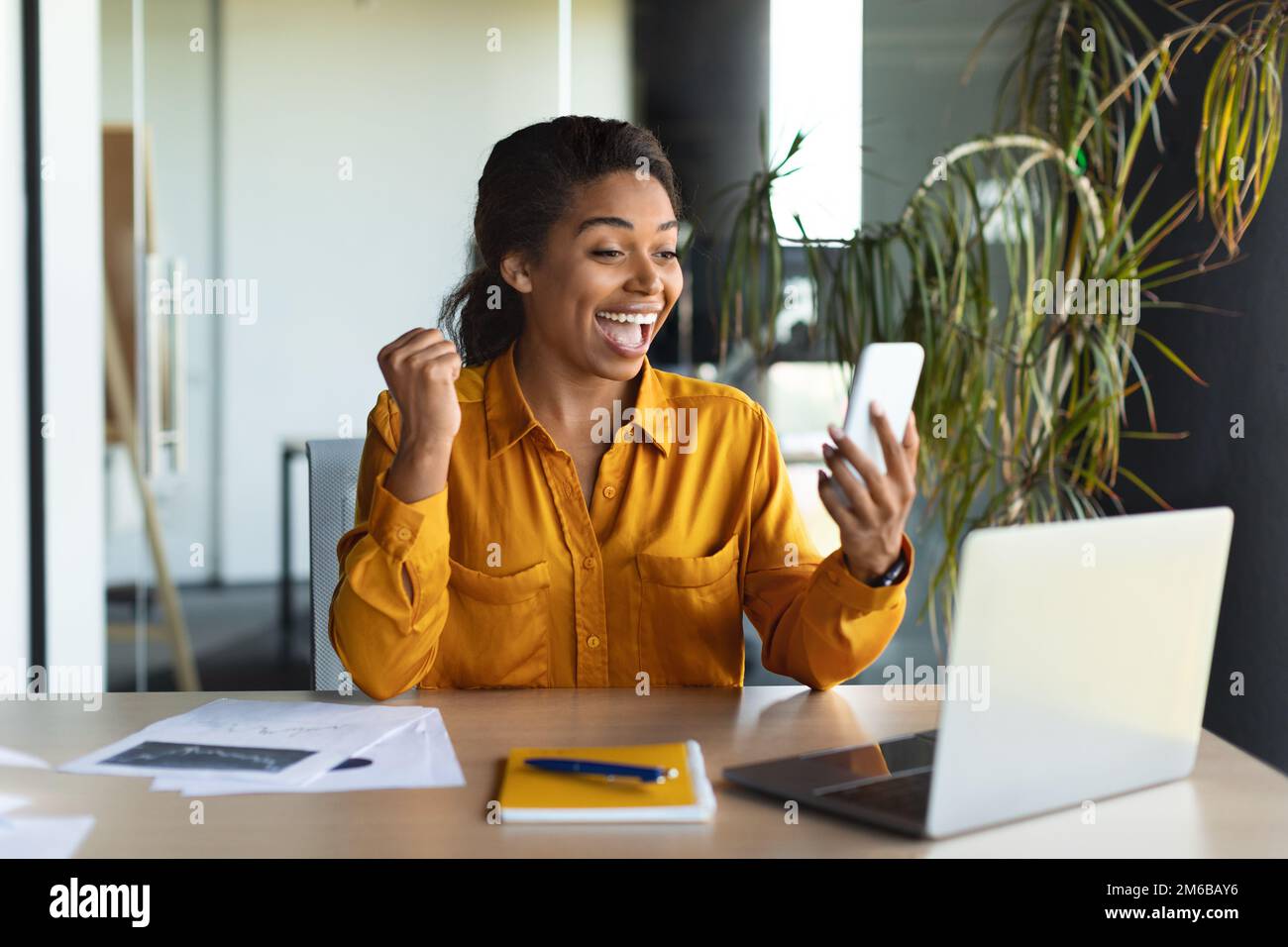 Femme noire excitée lisant un bon message sur un smartphone, travaillant sur un ordinateur portable dans un intérieur de bureau moderne Banque D'Images