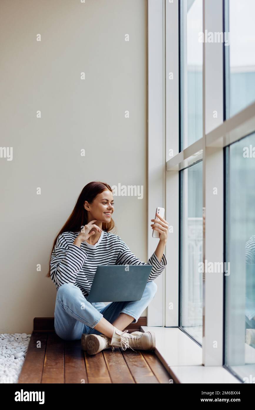 Femme indépendante assise avec ordinateur portable et téléphone à pleine fenêtre, appel vidéo, travail bonheur en ligne Banque D'Images