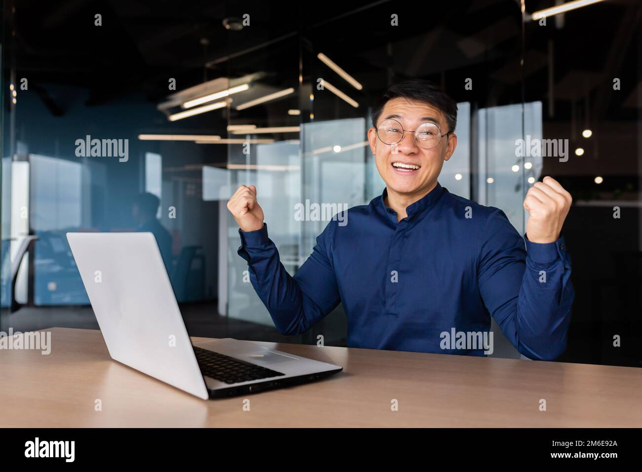 Homme d'affaires asiatique couronné de succès, portrait de l'homme au bureau en chemise décontractée, employé souriant et regardant l'appareil photo satisfait de la réussite en tenant le geste de la victoire main vers le haut. Banque D'Images