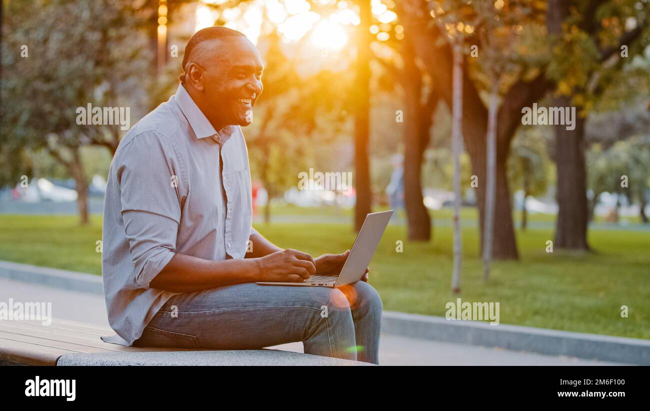 Joyeux insouciant senior africain homme américain assis à l'extérieur travail avec ordinateur portable jouer jeu en ligne ayant plaisir riant la saisie sur l'ordinateur. Maturité ethnique Banque D'Images