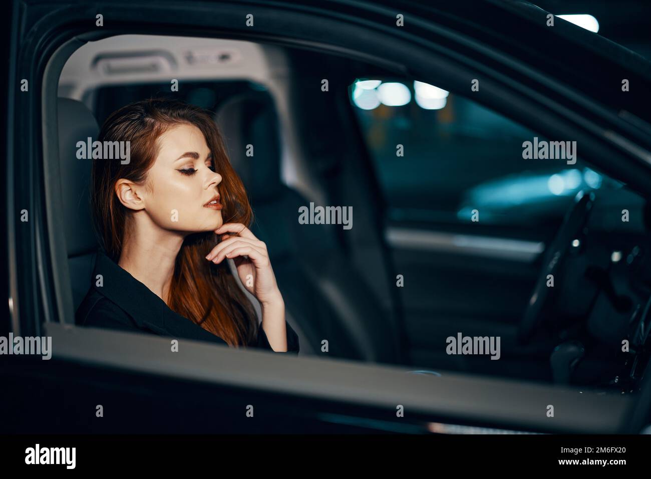 une femme élégante et luxueuse est assise dans une voiture noire la nuit, redressant ses cheveux longs et stylisés, fermant ses yeux avec plaisir Banque D'Images