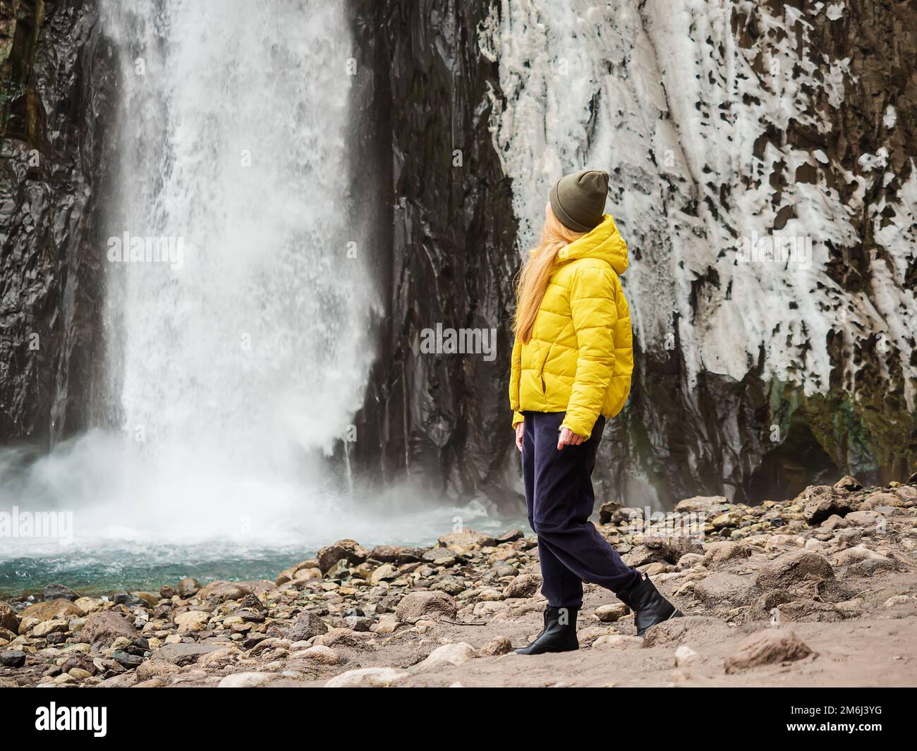Une femme dans une veste jaune regarde une cascade de glace orageux qui coule sur des rochers humides le jour froid de novembre. Banque D'Images