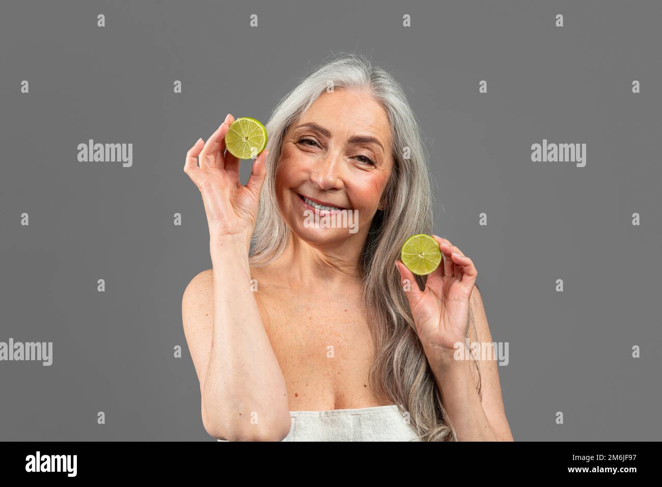 Vieille femme caucasienne souriante avec des morceaux de citrons gris, a l'amusement, isolé sur fond gris, studio Banque D'Images