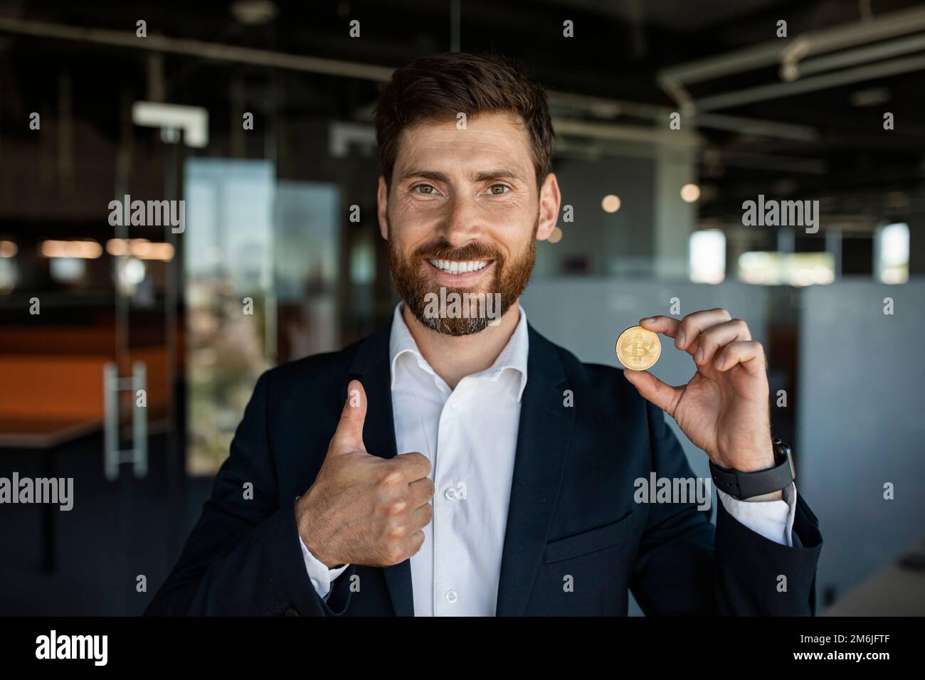 Un homme d'affaires heureux d'âge moyen montrant du bitcoin et se faisant le pouce dans un intérieur de bureau moderne, espace libre Banque D'Images