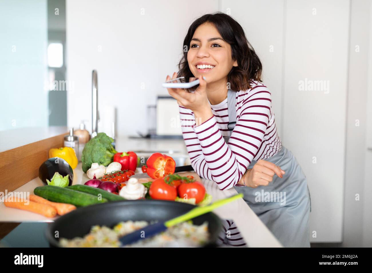 Jeune femme arabe positive enregistrant un message vocal pendant la cuisine Banque D'Images